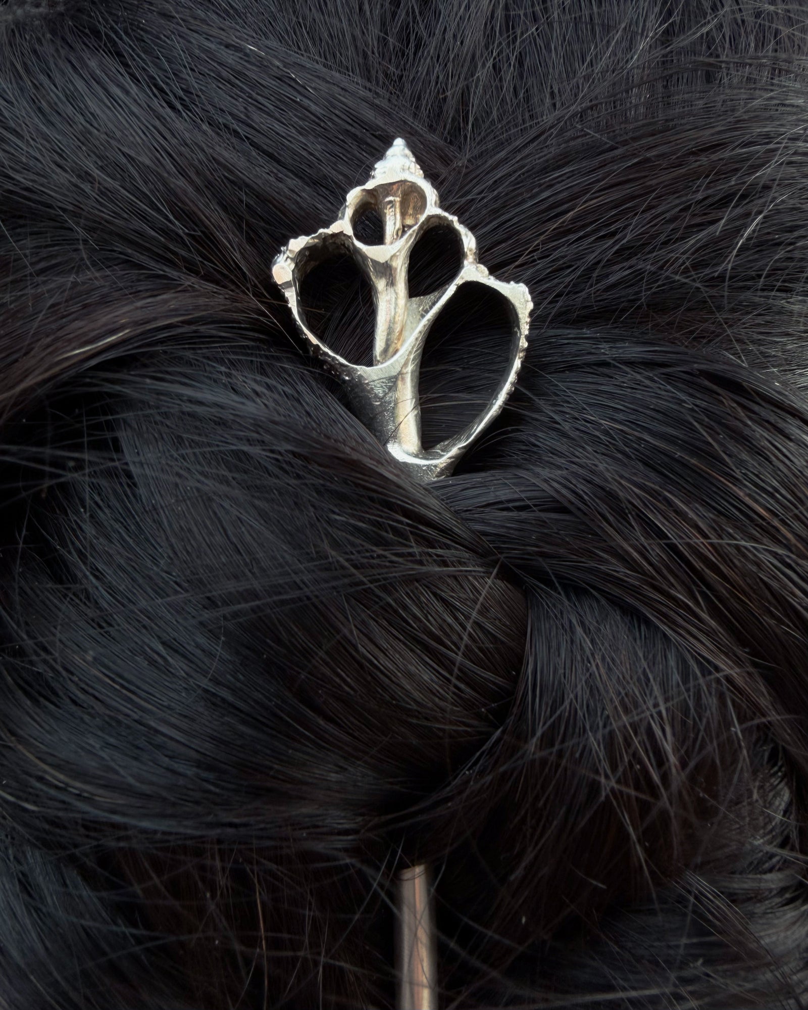 A silver metal hair accessory with a textured, organic shell silhouette, shown pinned onto a dark, wavy black hairpiece.